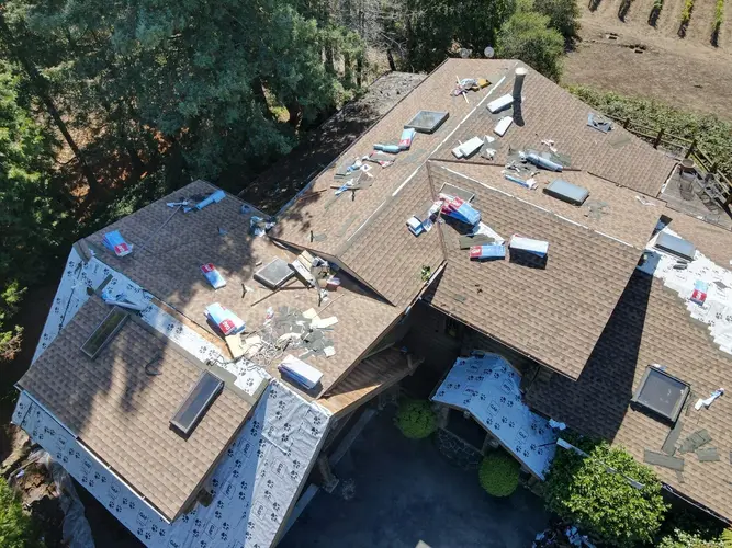 Roof repair and shingle installation in progress on a large residential home surrounded by trees and vegetation